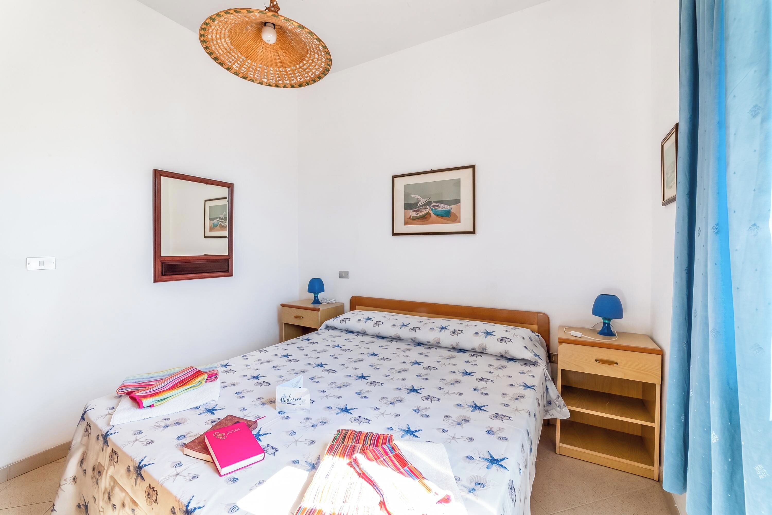 A bedroom with a bed, bedside tables, a hanging light fixture, a mirror, and a framed picture on the wall.
