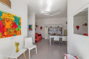 Ein modernes Wohnzimmer mit Essbereich, weißen Möbeln, einem Deckenventilator und einem farbenfrohen Gemälde an der Wand.