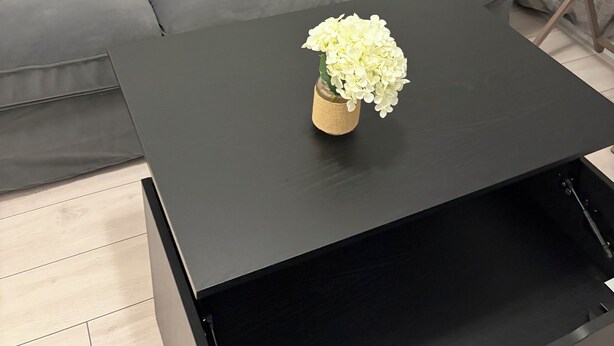 Een moderne woonkamer met een grijze bank, een zwarte salontafel met een vaas witte bloemen, en een klein houten bijzettafeltje.