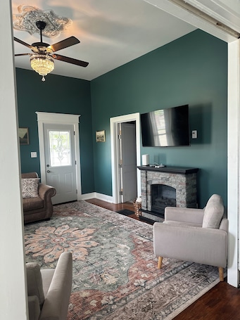 A living room with a fireplace, a television, a ceiling fan, and a patterned rug.. Verbena Executive Cottage