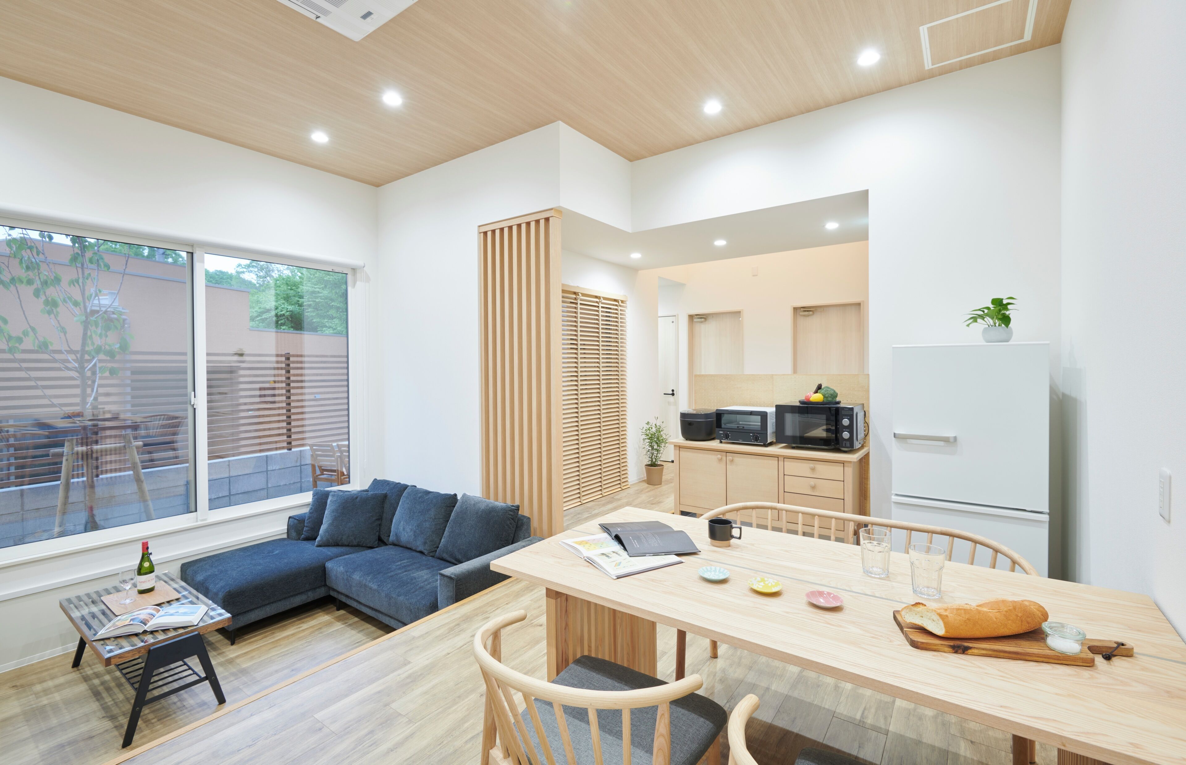 A modern living space with a sofa, dining table, chairs, kitchen area, and large windows.
