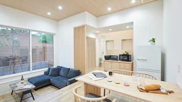 A modern living space with a sofa, dining table, chairs, kitchen area, and large windows.