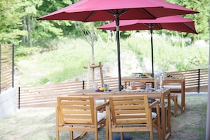 Een houten tafel met stoelen en een rode parasol, buiten opgesteld met een natuurlijke achtergrond.