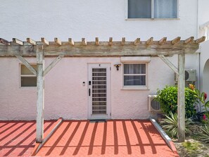 Ein Gebäude mit weißer Fassade, einer mit 'E' gekennzeichneten Tür und einem Fenster mit Vorhang, sowie einer Holzpergola.
