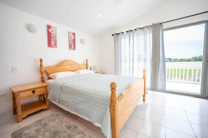 Ein Schlafzimmer mit einem hölzernen Bett, einem Nachttisch, einem flauschigen Teppich und Blick auf einen Balkon durch durchscheinende Vorhänge.