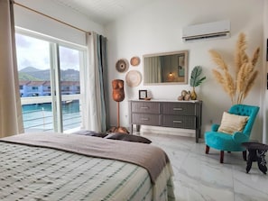 A bedroom with a large bed, a dresser, a chair, and a view of the sea. - Lazy Gecko Stylish with spacious deck (Saint John's)