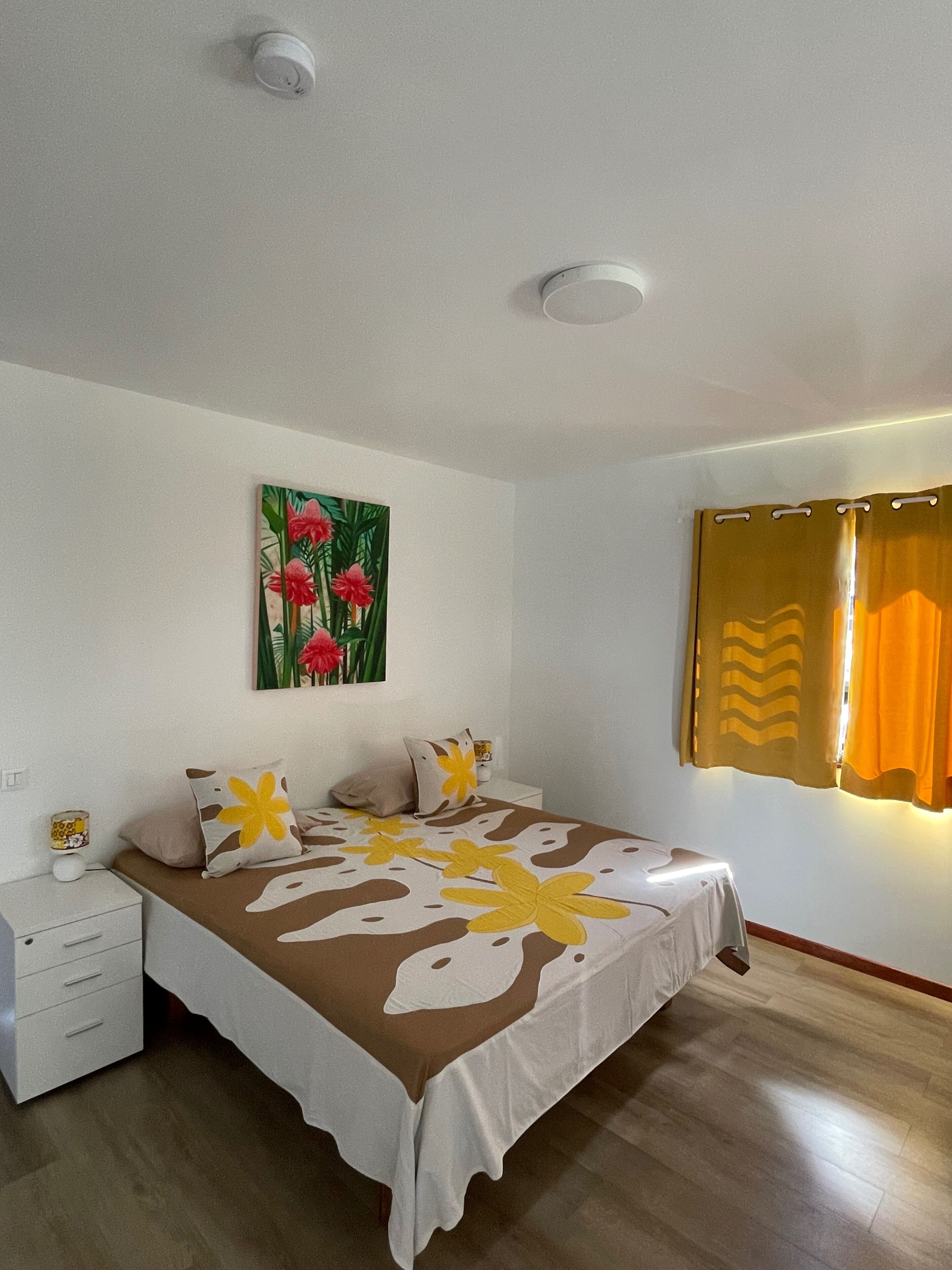 Ein Schlafzimmer mit einem Bett, einem Nachttisch, einem Wandbild und einem Fenster mit Vorhängen.