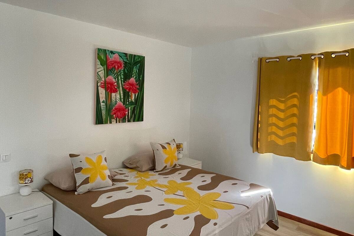 A bedroom with a bed, a nightstand, a wall-mounted painting, and a window with curtains.
