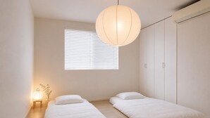 Ein schlicht eingerichtetes Schlafzimmer mit zwei Betten, einem Fenster mit Jalousien, einer Pendelleuchte und einem kleinen Nachttisch mit Lampe.