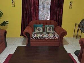 Uma sala de estar com sofá, almofadas estampadas, mesa de centro escura e um ventilador.