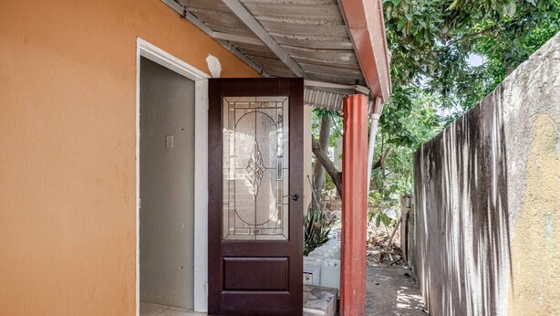 A house entrance with a dark wooden door, a tiled entrance, and a concrete pathway leading to a gated area. - Stylish 1BR Retreat 2 Queen Beds Quiet Upscale Area 15 mins to Town & Beaches (Spanish Town)