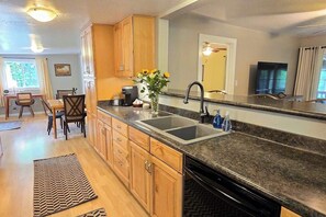 A modern kitchen with wooden cabinets, a granite countertop, a double sink, and a dining area with a chair and table. - Family Friendly Cottage w/ Meadowview, Hot Tub, Large Deck, WiFi & Private W/D (Avery)