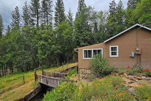 A cabin with a stone foundation, a wooden deck, and a small bridge over a stream. - Family Friendly Cottage w/ Meadowview, Hot Tub, Large Deck, WiFi & Private W/D (Avery)