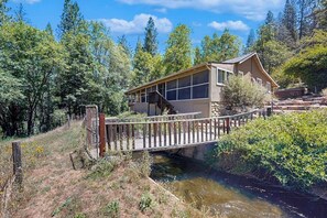 A cabin with a wooden deck, a small bridge, and a natural setting with trees and a stream. - Family Friendly Cabin w/ Meadow View, Hot Tub, Large Deck, WiFi & Private W/D (Avery)