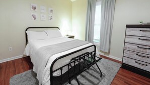 Um quarto com cama, cômoda, mesa de cabeceira, janela com cortinas e obras de arte nas paredes.