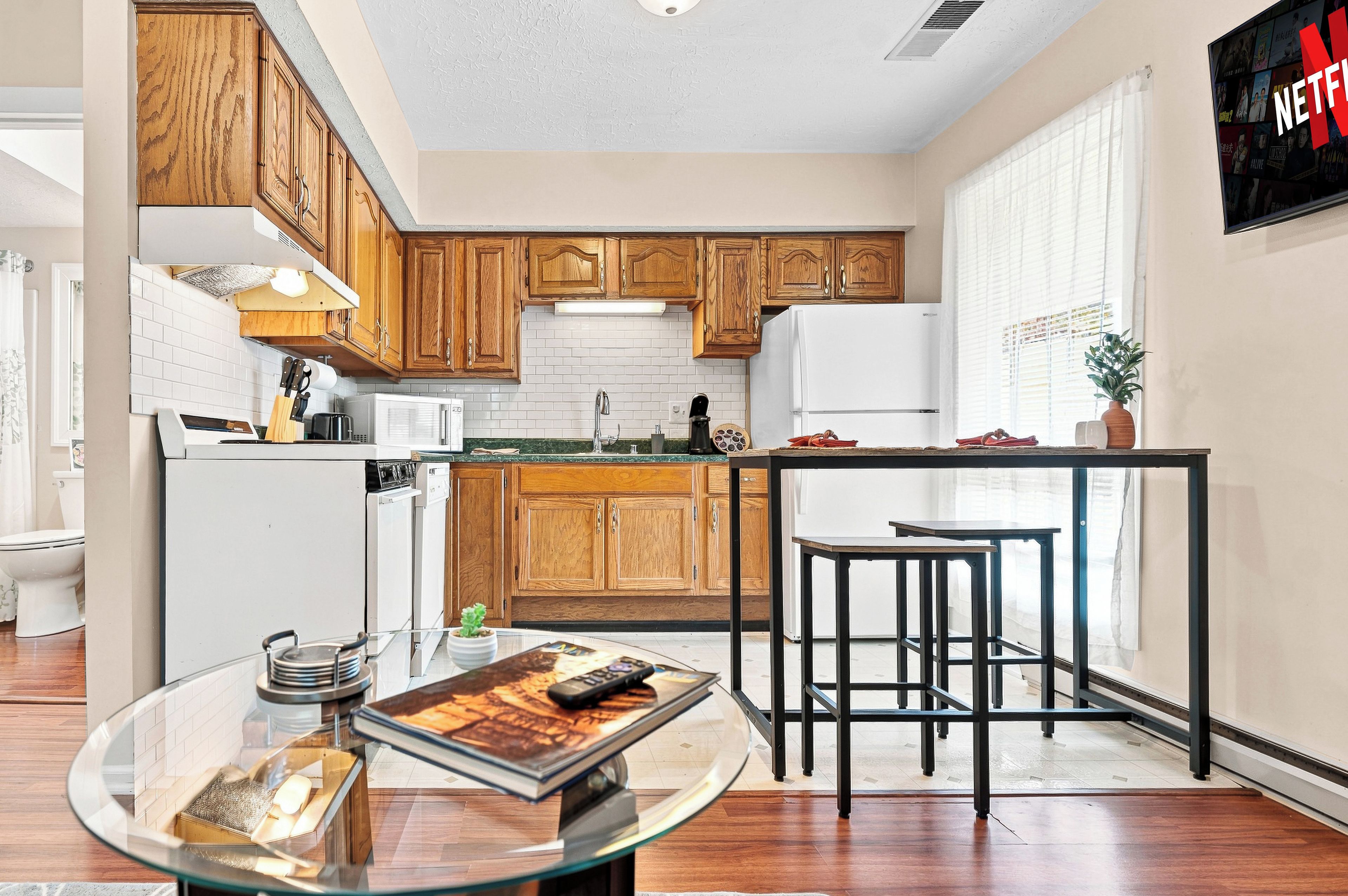 A modern kitchen with wooden cabinets, a white refrigerator, a coffee machine, and a glass table with a book.
