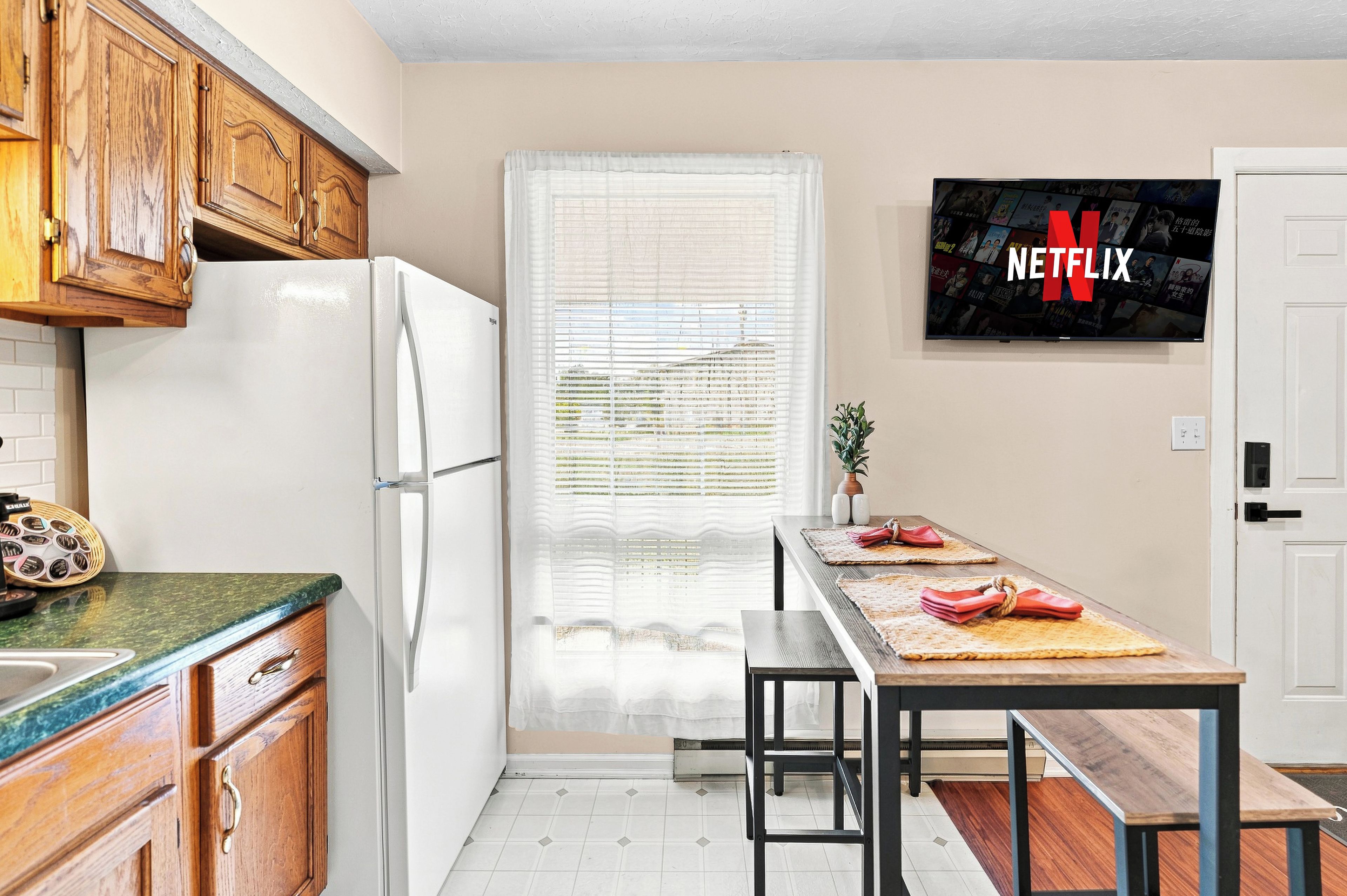 Una cocina con armarios de madera, un refrigerador blanco, una mesa de comedor con bancos y un televisor instalado en la pared que muestra el logotipo de Netflix.