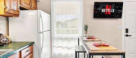 Una cocina con armarios de madera, un refrigerador blanco, una mesa de comedor con bancos y un televisor instalado en la pared que muestra el logotipo de Netflix.