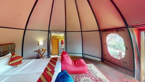 Una tienda de campaña con una cama, almohadas, una mesita con una lámpara y vistas a través de un ventanal redondo.