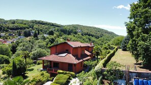 A two-story house with a red roof, surrounded by lush greenery and hills in the background.