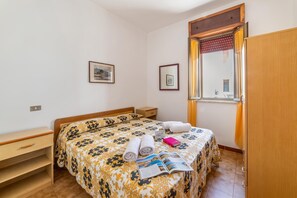 Ein Schlafzimmer mit einem Bett, einem Nachttisch, einer hölzernen Kommode, einem Fenster mit Vorhängen und einem an der Wand befestigten Regal.