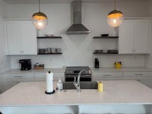 Una cocina con armarios blancos, una isla central con un fregadero, una estufa y dos luces colgantes.