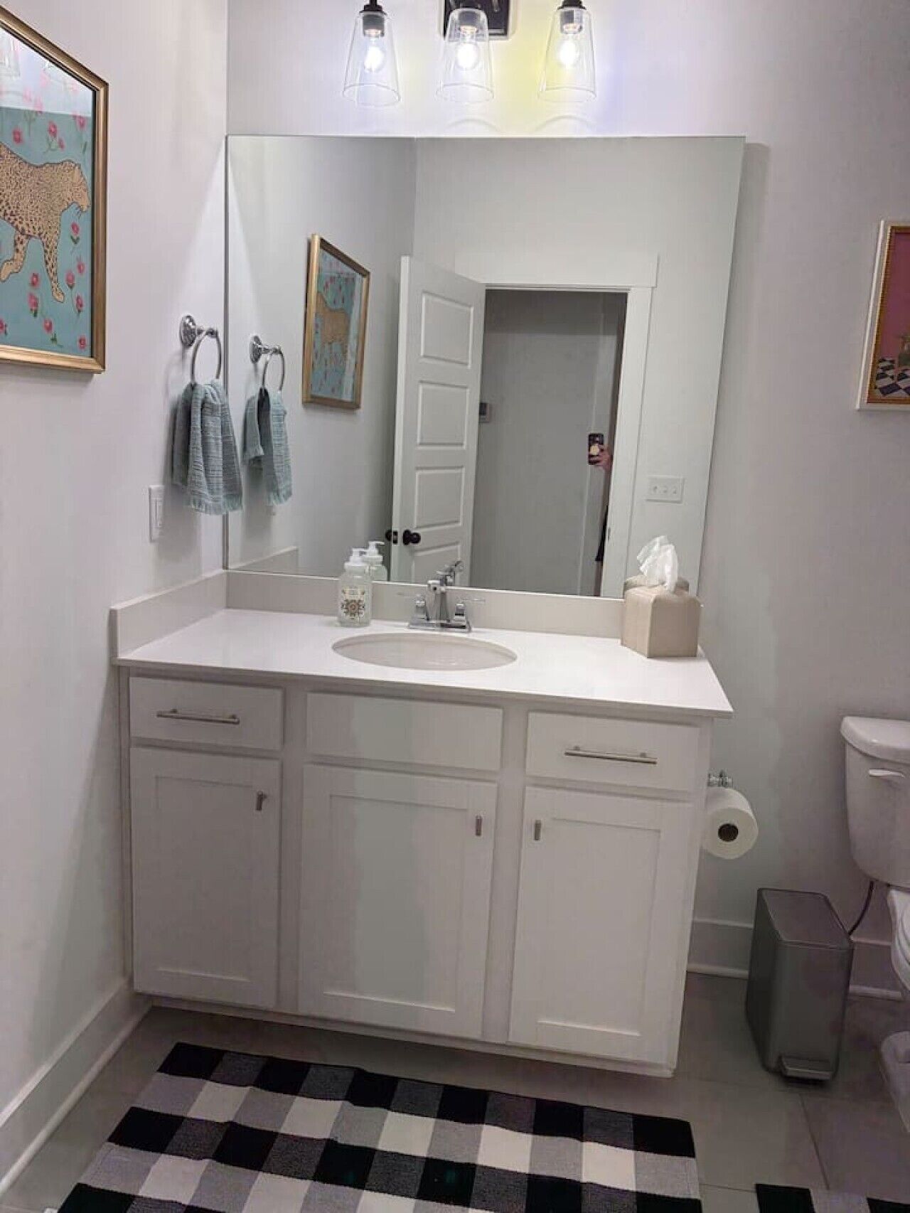 A bathroom with a white vanity, a large mirror, a toilet, and a black and white checkered rug.