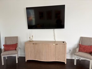Ein Wohnzimmer mit einem an der Wand montierten Flachbildfernseher, einer Holzkassette mit Schubladen, zwei Sesseln mit roten Kissen und einem dunklen Holzboden.