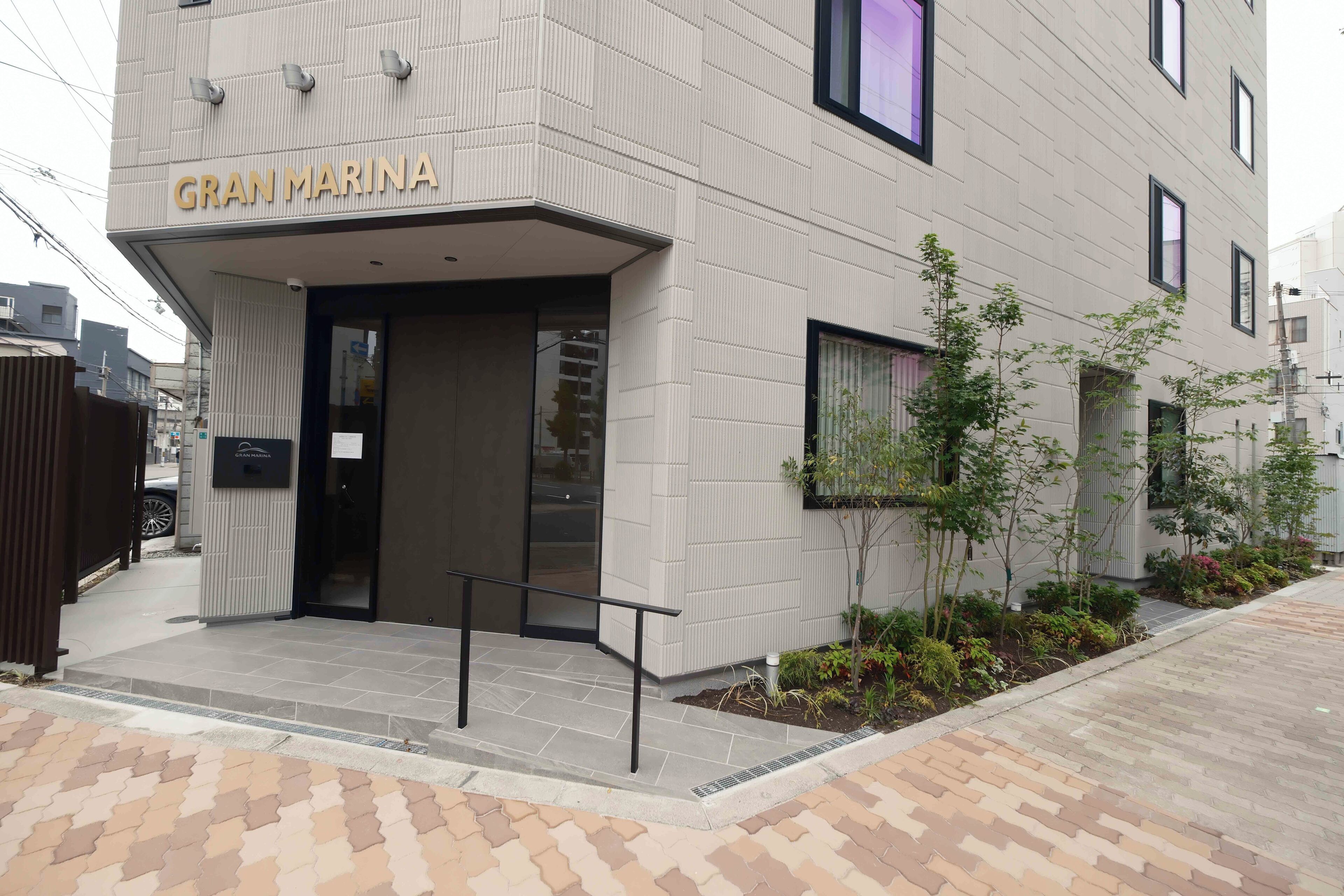 A building with a sign reading 'GRAN MARINA' at the entrance, a tiled pathway, and a small landscaped area with plants.
