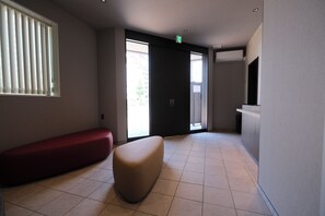 Un vestíbulo de hotel con un banco rojo, un reposapiés color beige, un mostrador de recepción y un amplio ventanal corredera.