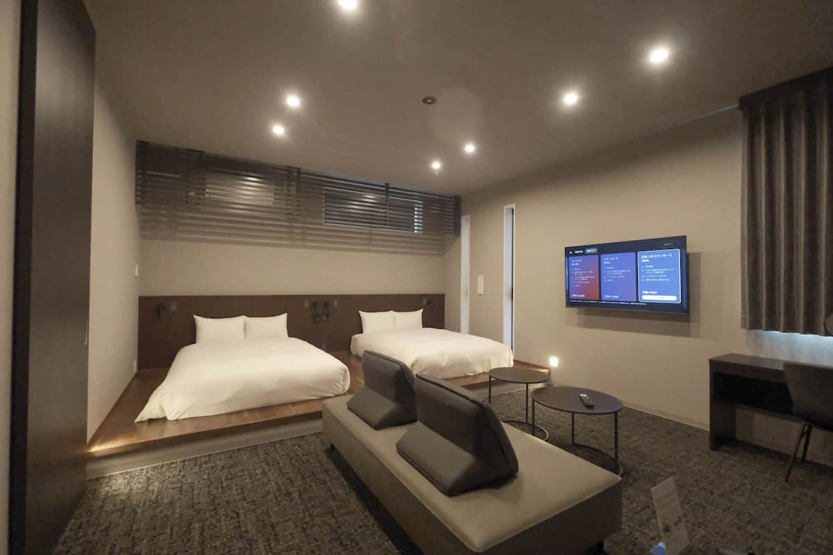 A hotel room with two beds, a sofa, a desk, and a TV displaying room information.