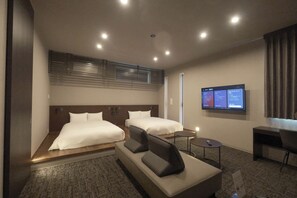 Quarto de hotel com duas camas, um sofá, uma escrivaninha e uma TV exibindo informações do quarto.