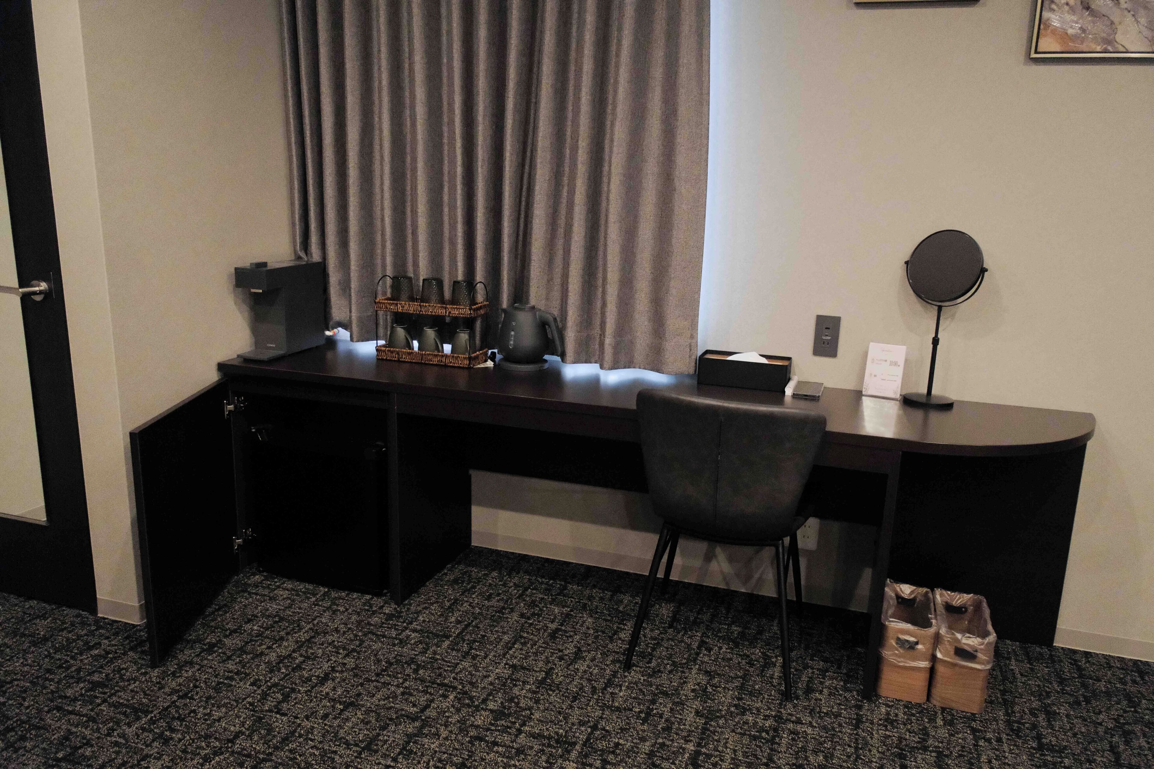 A hotel room with a desk, a chair, a coffee maker, a kettle, a trash bin, and a wall-mounted mirror.