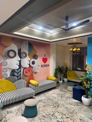 A modern living room with a ceiling fan, a sofa with yellow cushions, a blue ottoman, and a wall mural. - HOTEL ELITE CHOICE (Ajmer)