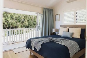 Quarto com cama, mesa de cabeceira, abajur, janela com persianas e vista para o verde lá fora.