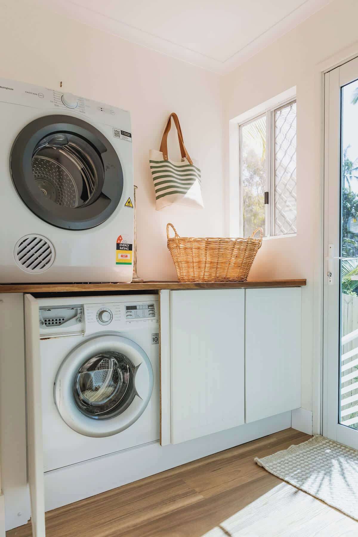 A laundry room with a front-loading washer and dryer, a wicker basket on the counter, and a window with a view of greenery.