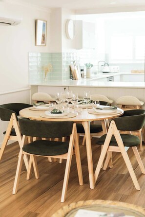 Uma sala de jantar com mesa redonda de madeira, cadeiras estofadas em verde escuro e uma cozinha ao fundo.