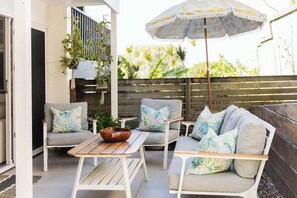 A patio with a wooden table, two white sofas with patterned cushions, and a large umbrella. - Hidden Sunshine Beach Gem • Steps to the Beach! (Sunshine Beach)