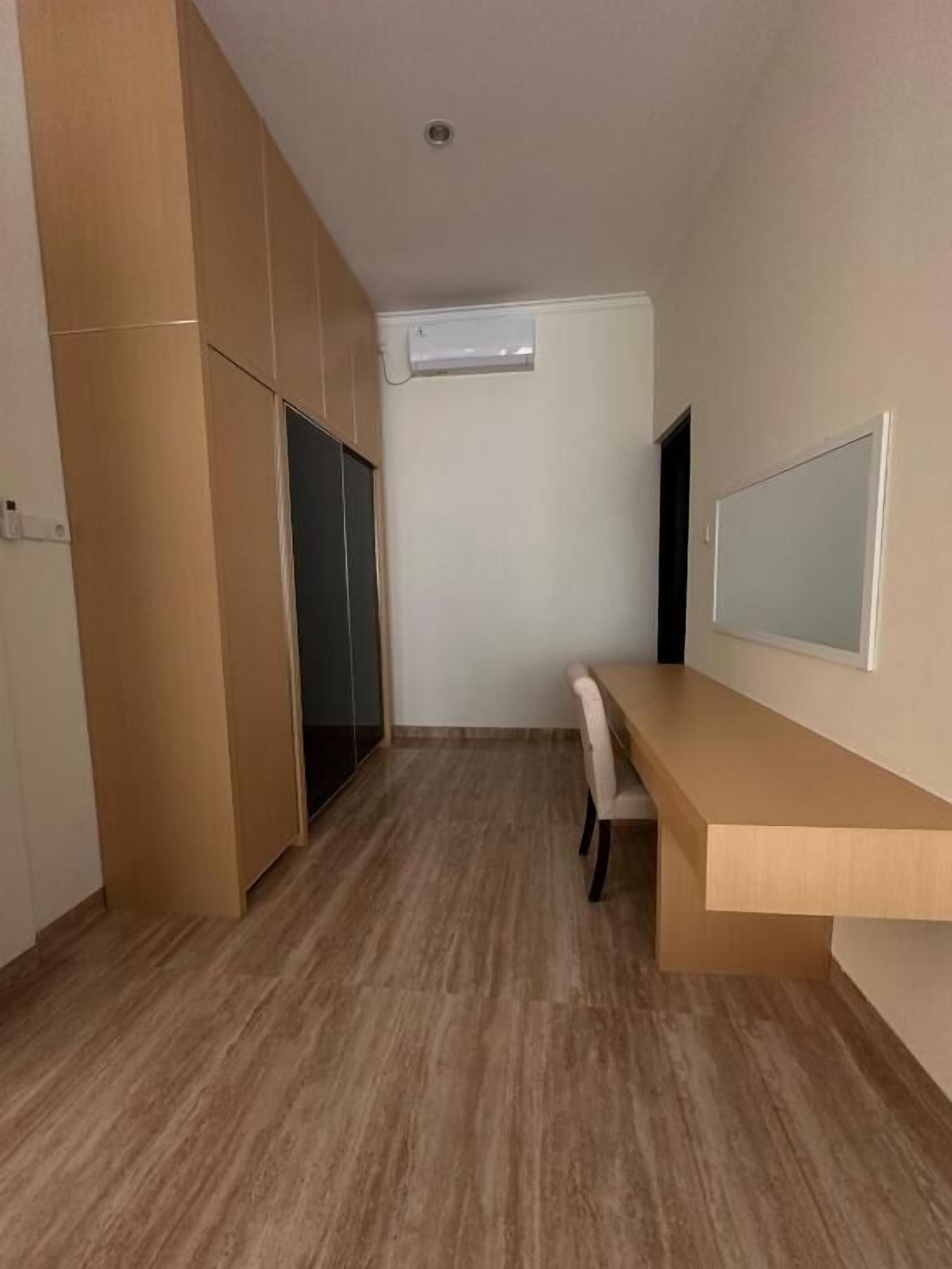 A room with a wooden floor, a desk with a chair, a white wall, and a ceiling vent.