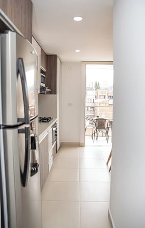 Cozinha moderna com eletrodomésticos de aço inoxidável, área de jantar com mesa e cadeiras, e vista para edifícios através de uma grande janela.