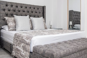 Quarto com cabeceira estofada, cama grande com colcha estampada, várias almofadas, mesa de cabeceira com abajur e um banco ao pé da cama.