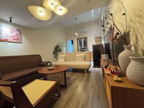 Ein Wohnzimmer mit braunem Sofa, einem runden Couchtisch und einem beigen Sofa. Es gibt Korbstühle, eine dekorative Vase und eine wandmontierte Klimaanlage.