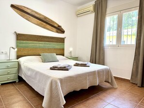 Quarto com cabeceira de madeira, cama com colcha branca, duas mesas de cabeceira com abajures e uma janela com cortinas.