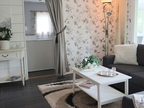Una sala de estar con una mesa de centro blanca, un sofá gris, un papel tapiz estampado, una lámpara de pie y una planta pequeña sobre una mesita auxiliar.