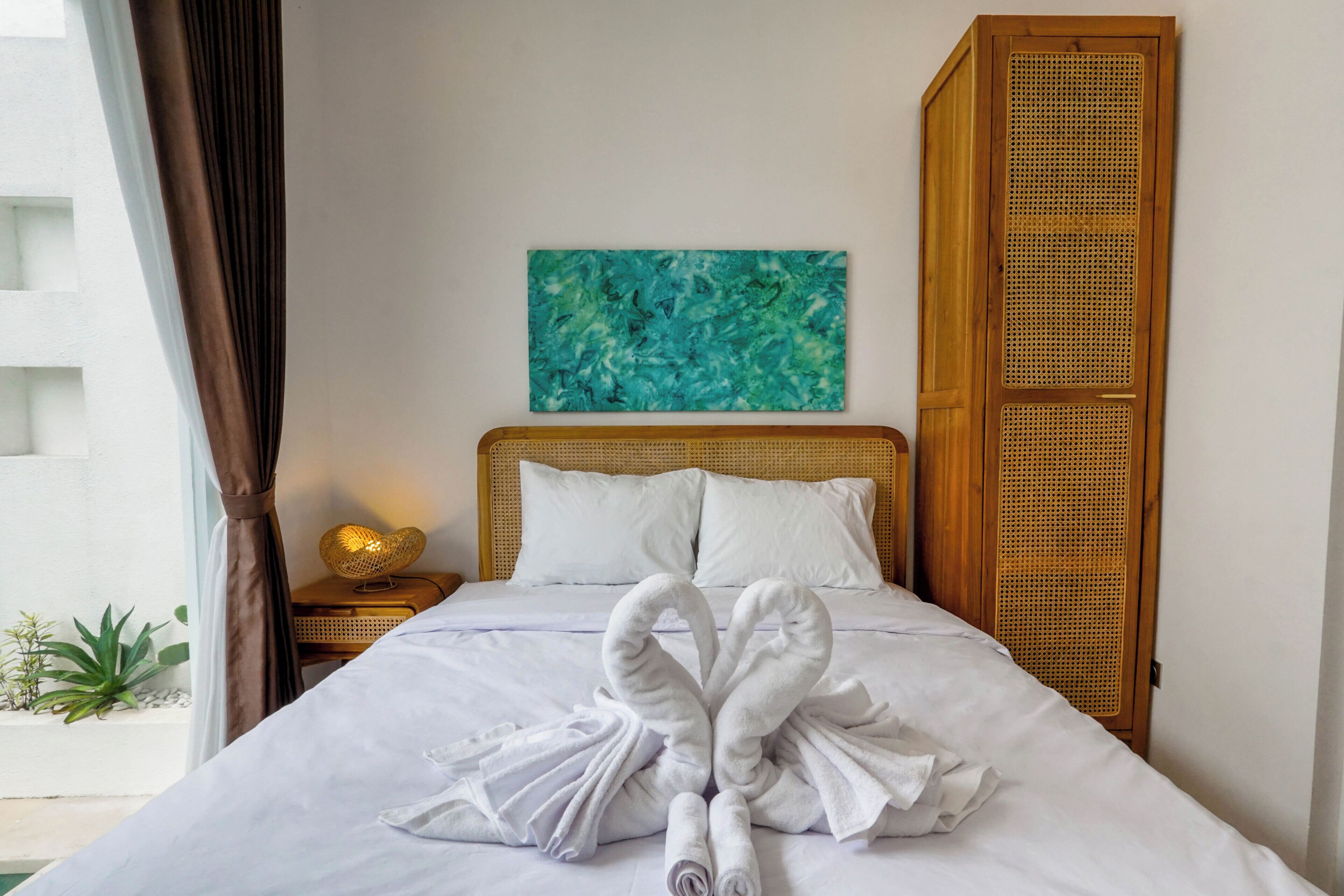 Une chambre d’hôtel avec un lit, une tête de lit en rotin, une œuvre d’art accrochée au mur et une table de chevet agrémentée d’un objet décoratif.