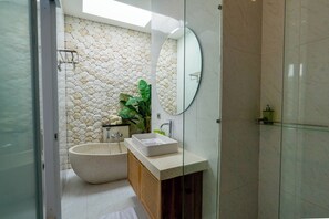 A bathroom with a stone wall, a freestanding bathtub, a mirror, and a sink with a wooden cabinet. - Family 2 Bedroom Villa by Seminyak - B13 (Kota Denpasar)