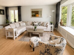 A living room with a sofa, a coffee table, a side table, and a chair. There is a window with curtains, a floor lamp, and a mirror on the wall.