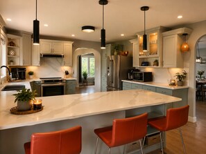 A modern kitchen with a central island, stainless steel appliances, pendant lighting, and a view to a dining area.