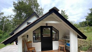 Una pequeña cabaña de madera con techo inclinado, un porche con dos sillas de madera y una puerta con ventana.