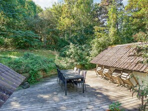 A wooden deck with a table and chairs, surrounded by dense greenery and a rustic building with a tiled roof. - 4 Star Holiday Home in Sejero (Sejero)